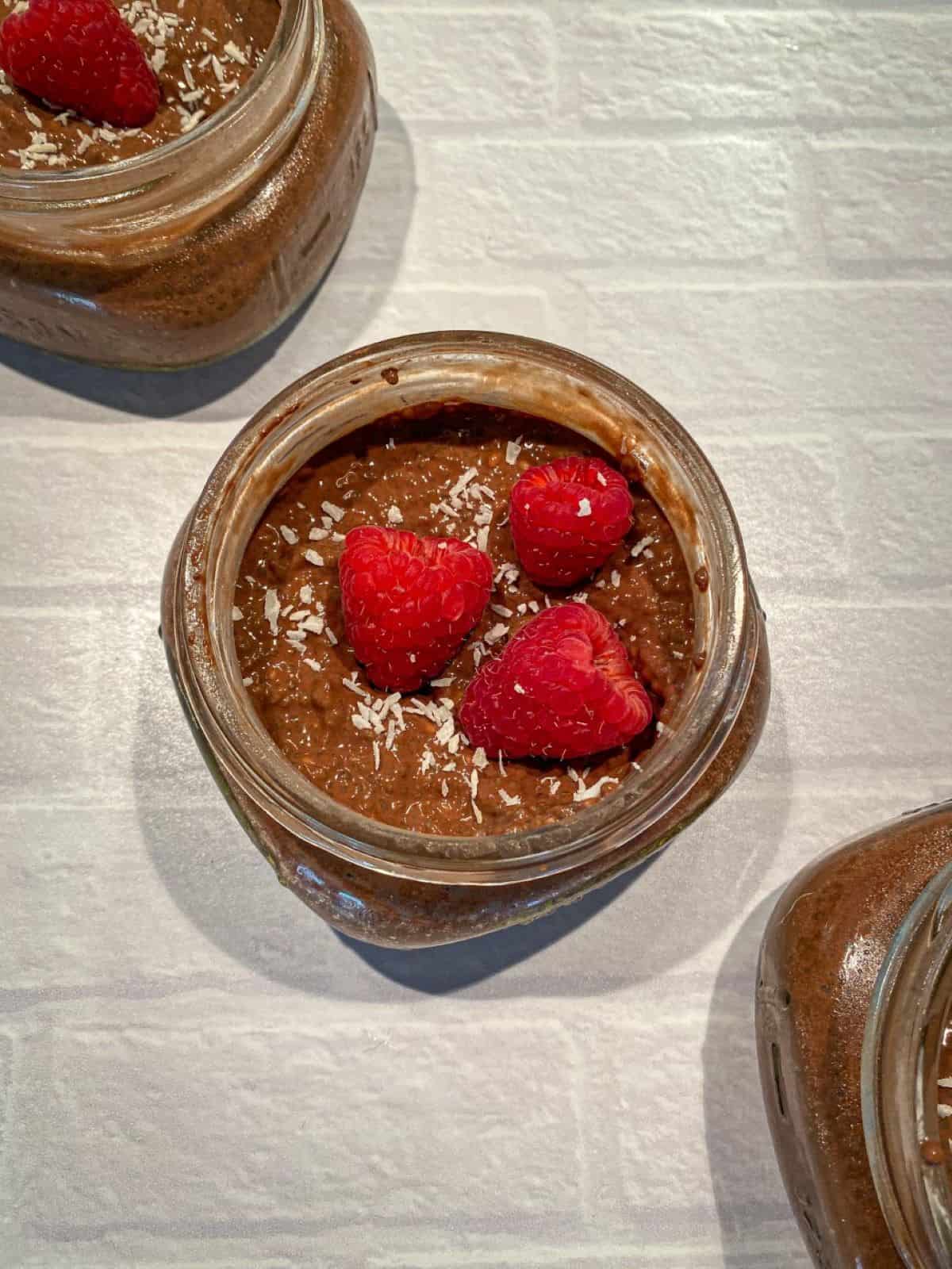 Three mason jars full of chocolate chia pudding topped with raspberries and coconut flakes.