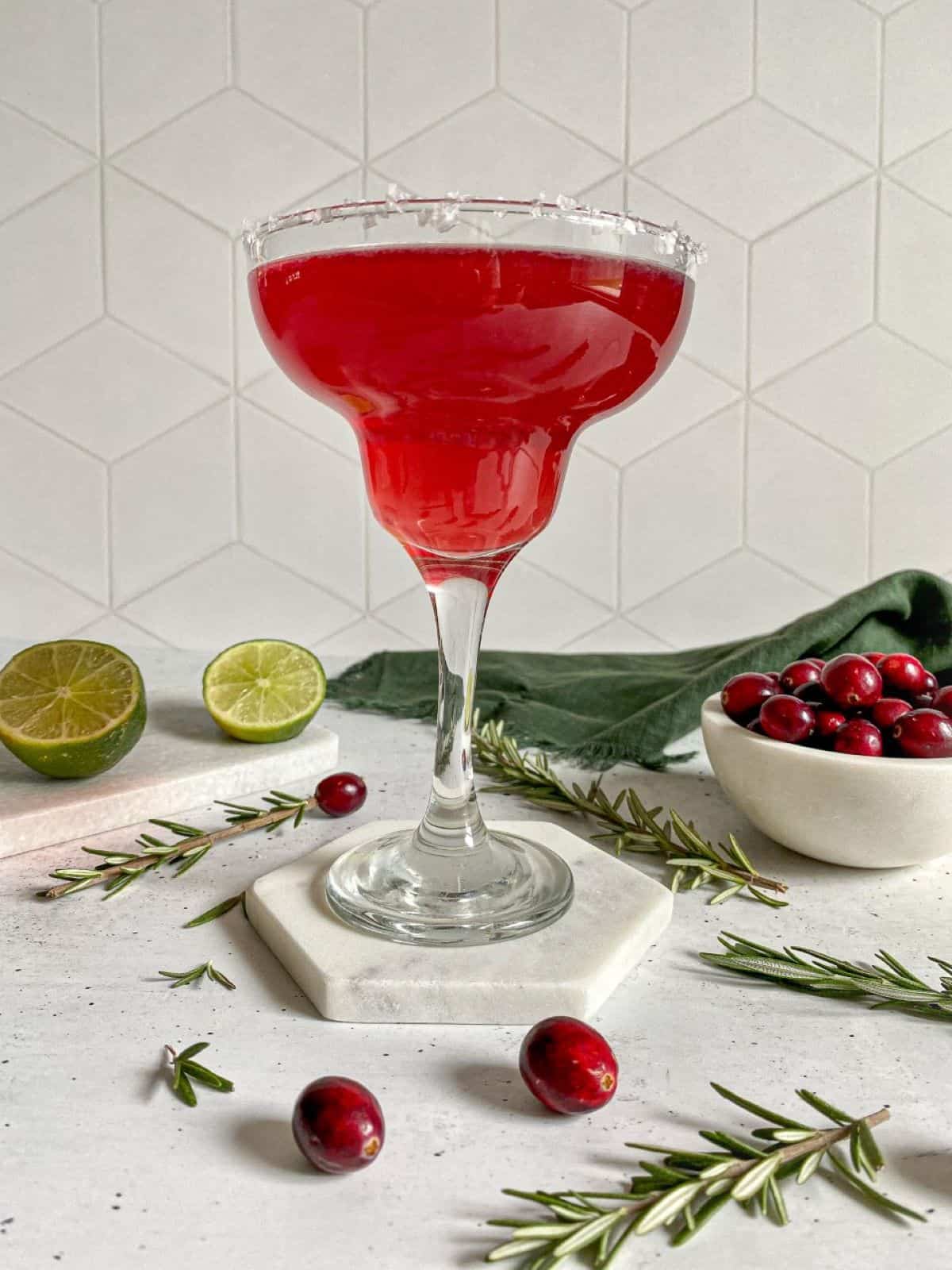 A cranberry margarita in a glass with a salt rim surrounded by fresh cranberries and limes.