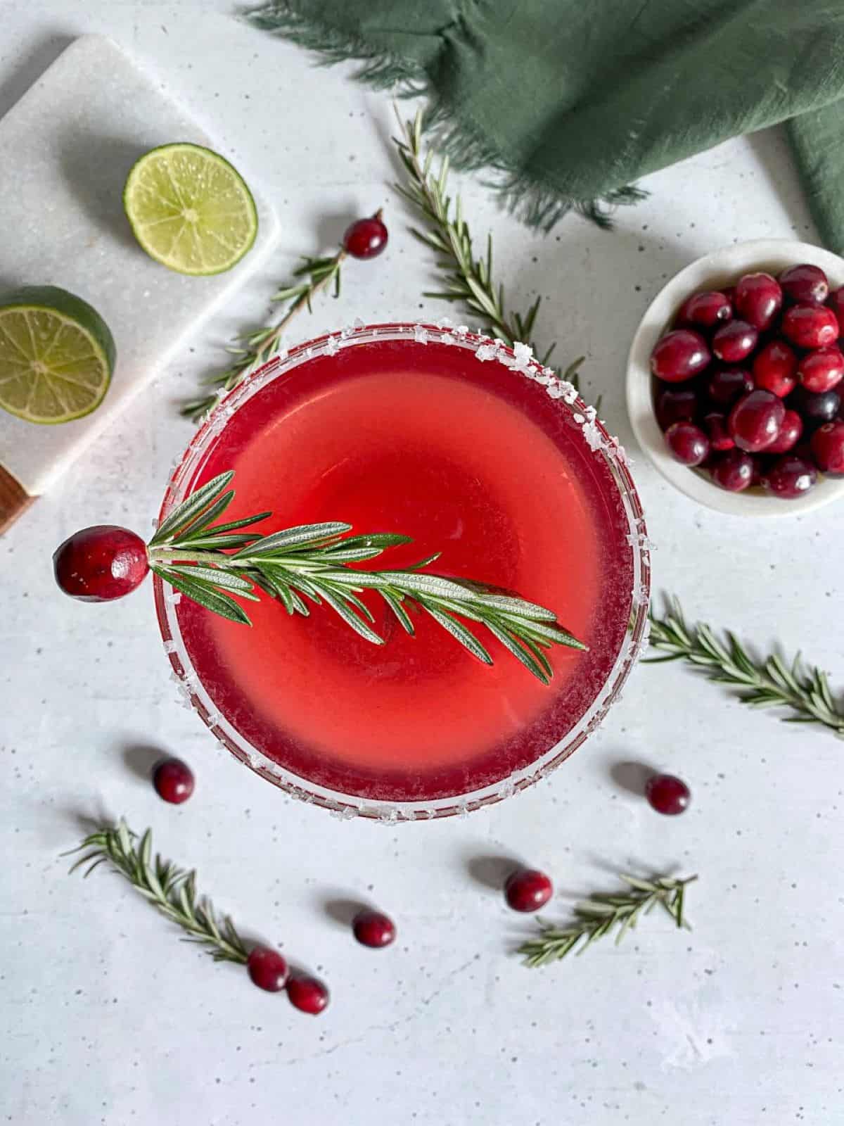 A red cocktail in a glass with a salted rim. A garnish of rosemary and a cranberry are on top of the drink.