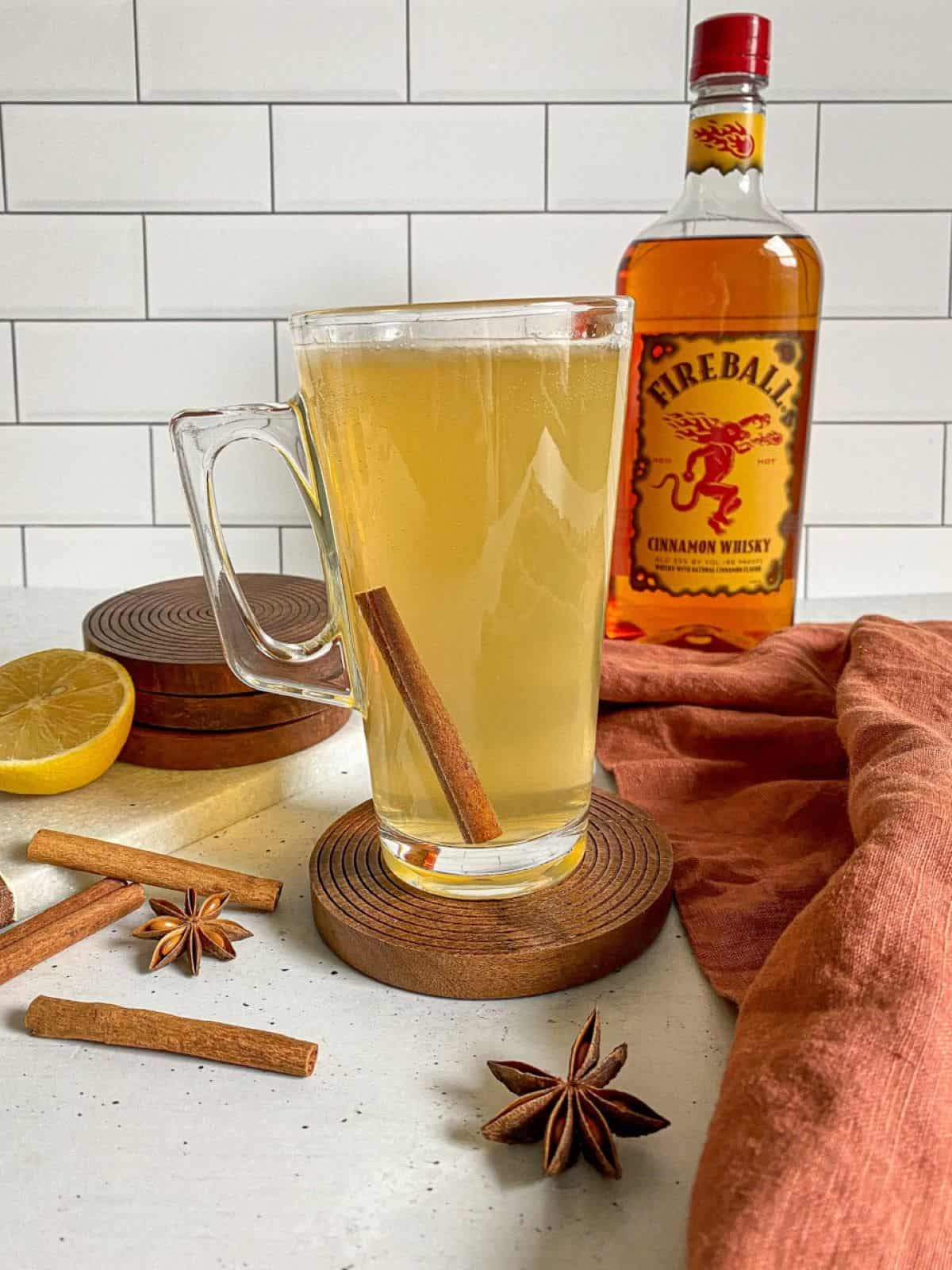 A tall clear mug is filled with Fireball Hot Toddy. The drink is surrounded by a bottle of whiskey, sliced lemon, and cinnamon sticks.