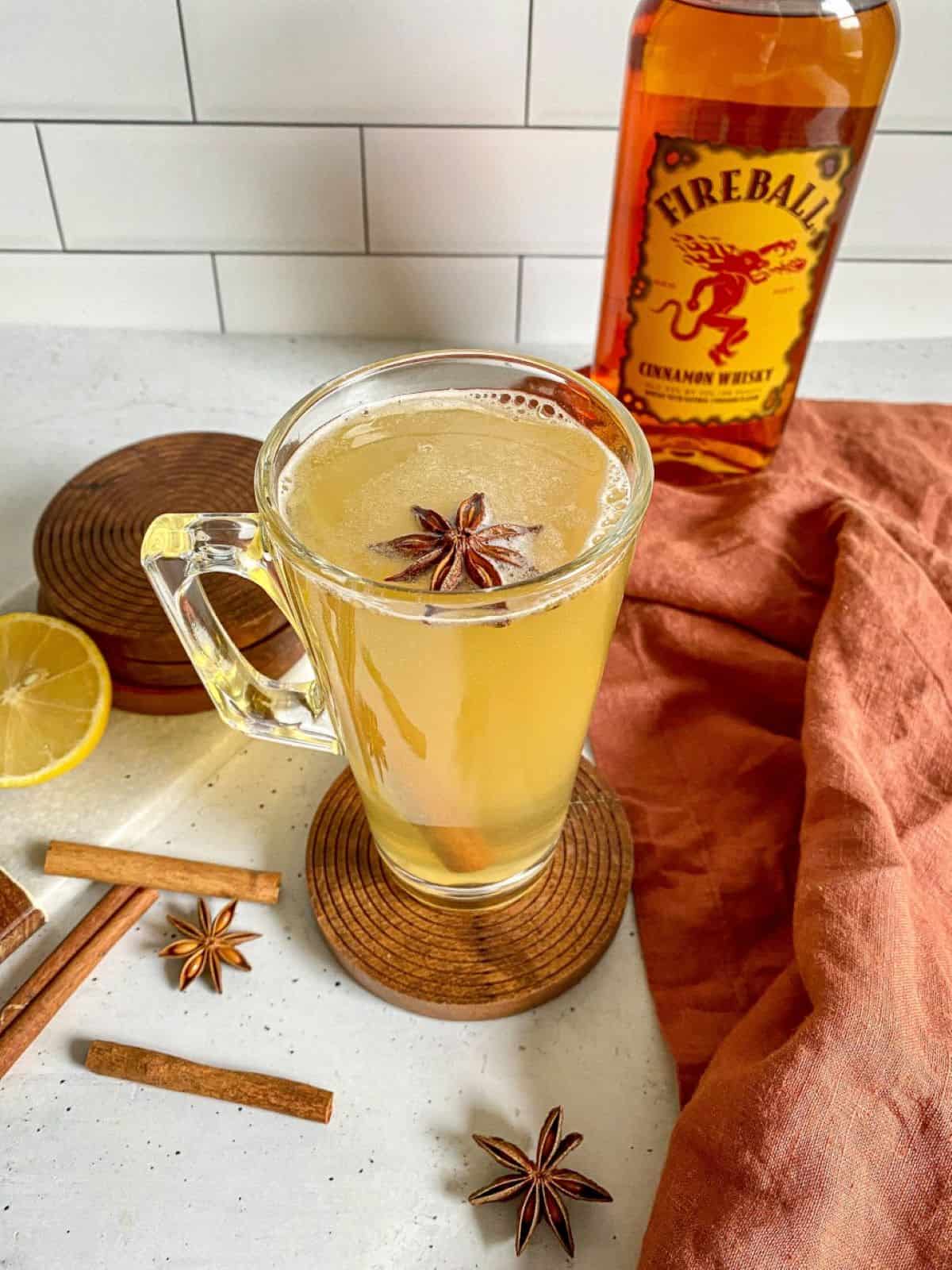 Hot Toddy in a clear mug surrounded by sliced lemon and cinnamon sticks. A bottle of Fireball whisky is next to the cocktail.