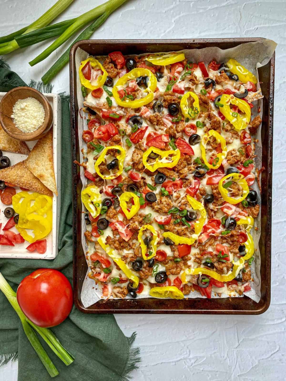Italian Nachos on a sheet pan topped with sausage, alfredo sauce, cheese, tomatoes, olives, and peppers. A tray with extra toppings and grated cheese is next to the pan.