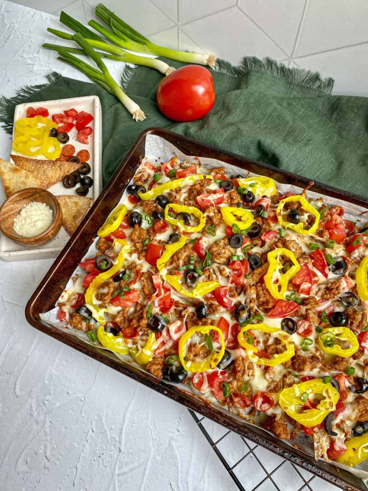 Sheet pan nachos topped with sausage, alfredo sauce, cheese, tomatoes, olives, and peppers. A tray with extra toppings, green onions, and grated cheese is next to the pan.