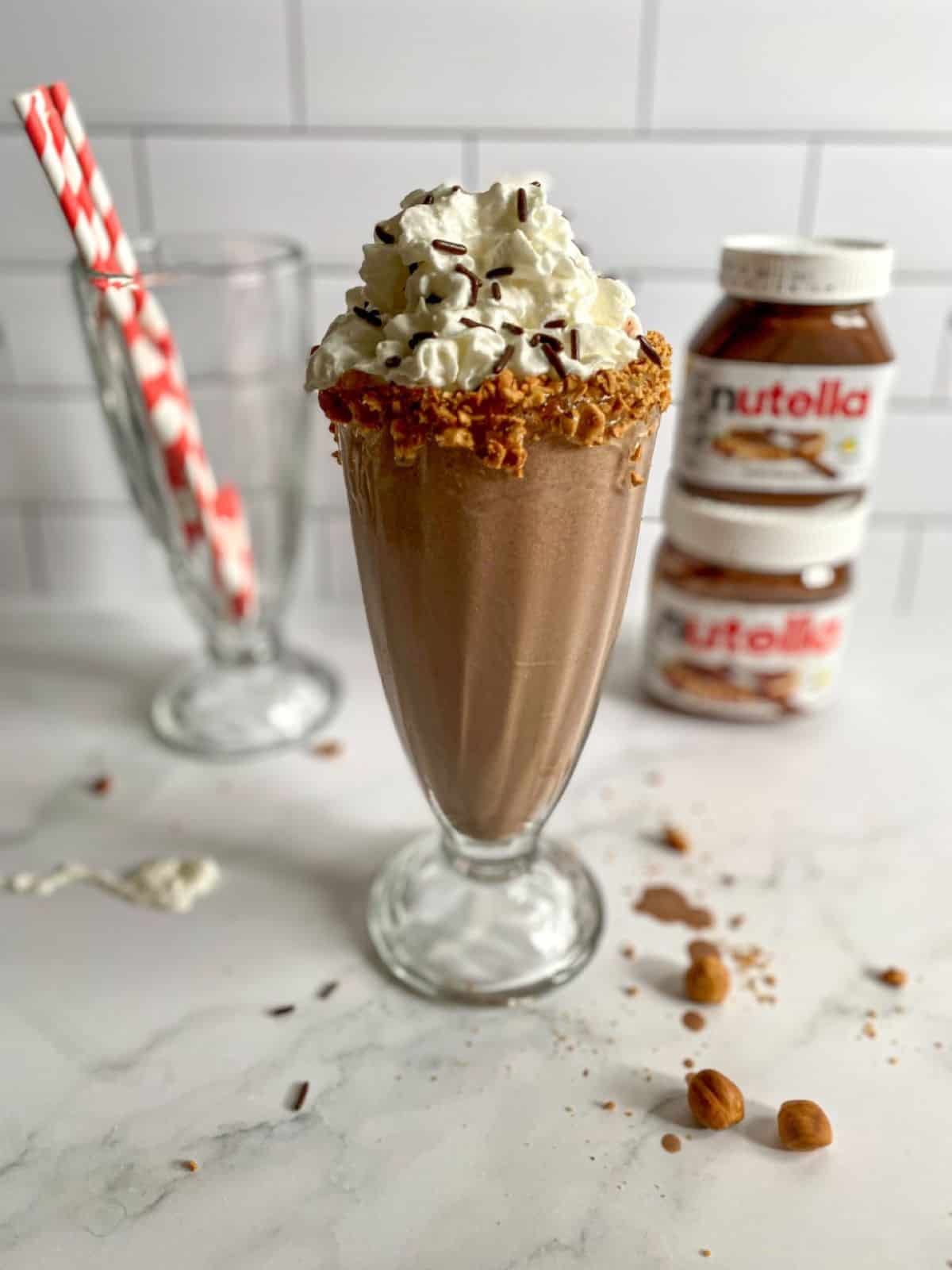 Nutella milkshake in a clear glass topped with whipped cream and sprinkles in a tall old fashioned milkshake glass. Jars of Nutella and red straws are in the background.