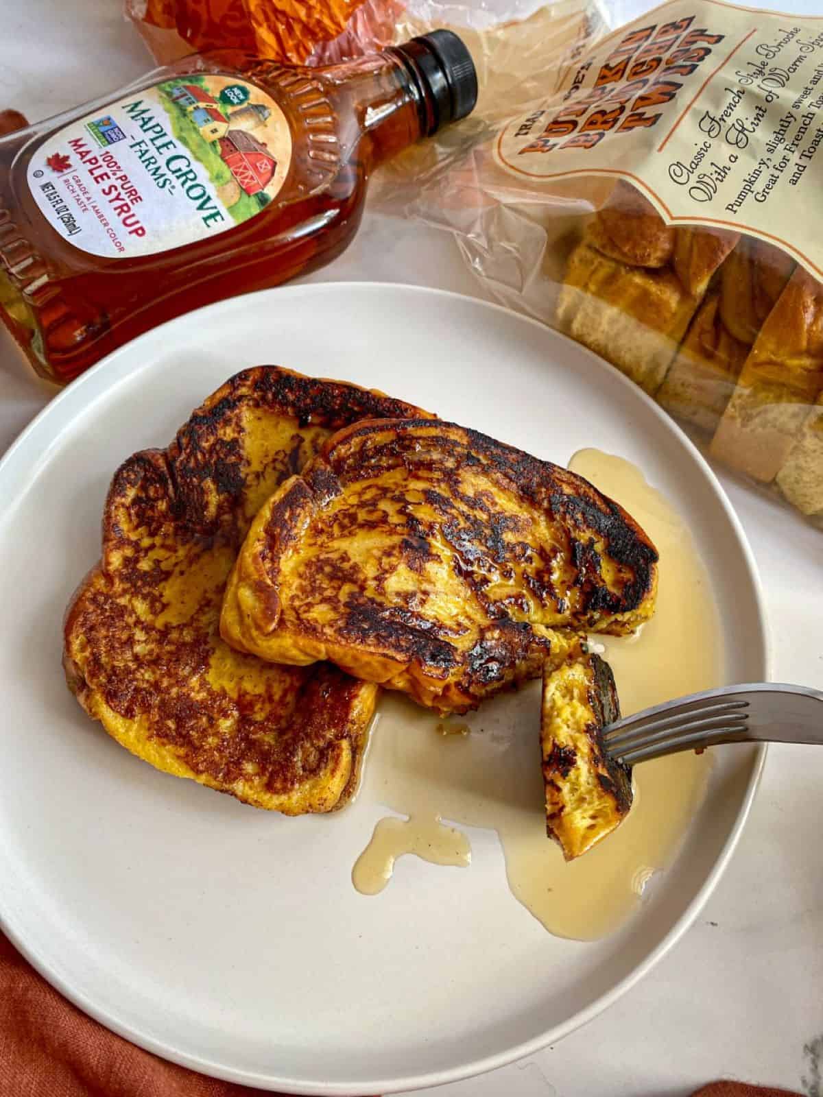 Two slices of brioche French toast are on a white plate with a fork cutting into one of the slices showing the soft inside. A glass bottle of maple syrup and a loaf of bread are next to the plate.