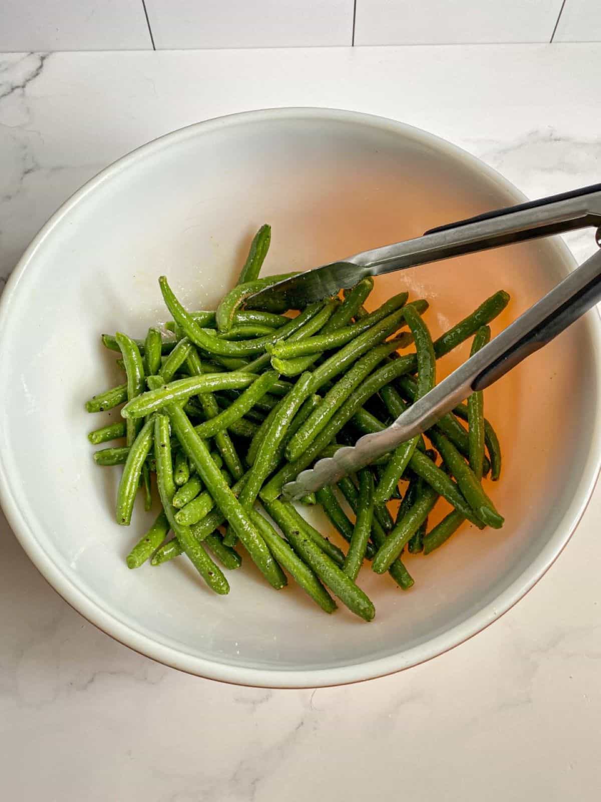Seasoned green beans in a mixing bowl with tongs.