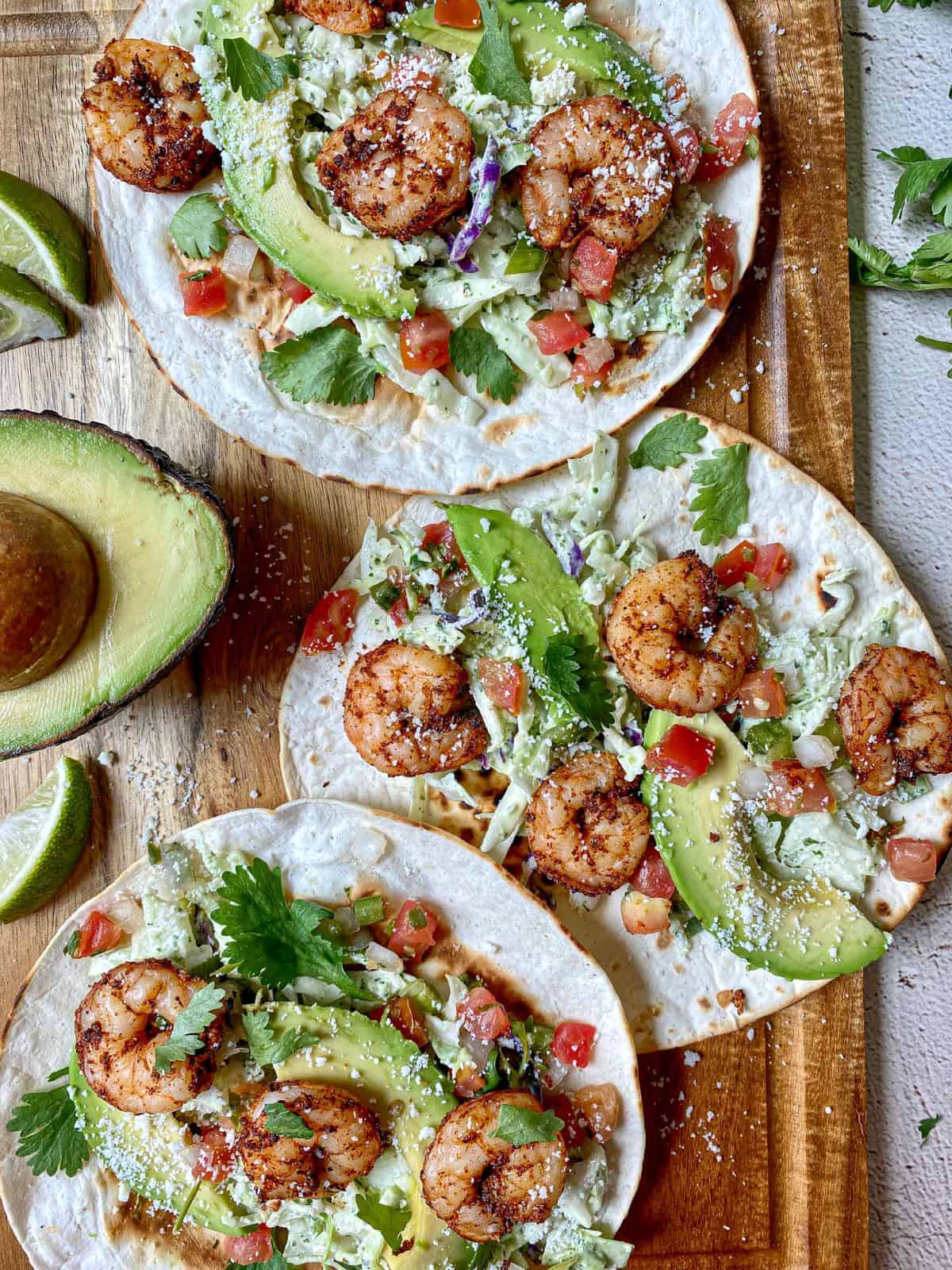 Three shrimp tacos with slaw, avocado, cilantro, and creamy jalapeno salsa are open face on a cutting board. Lime wedges and an avocado are on the side. 