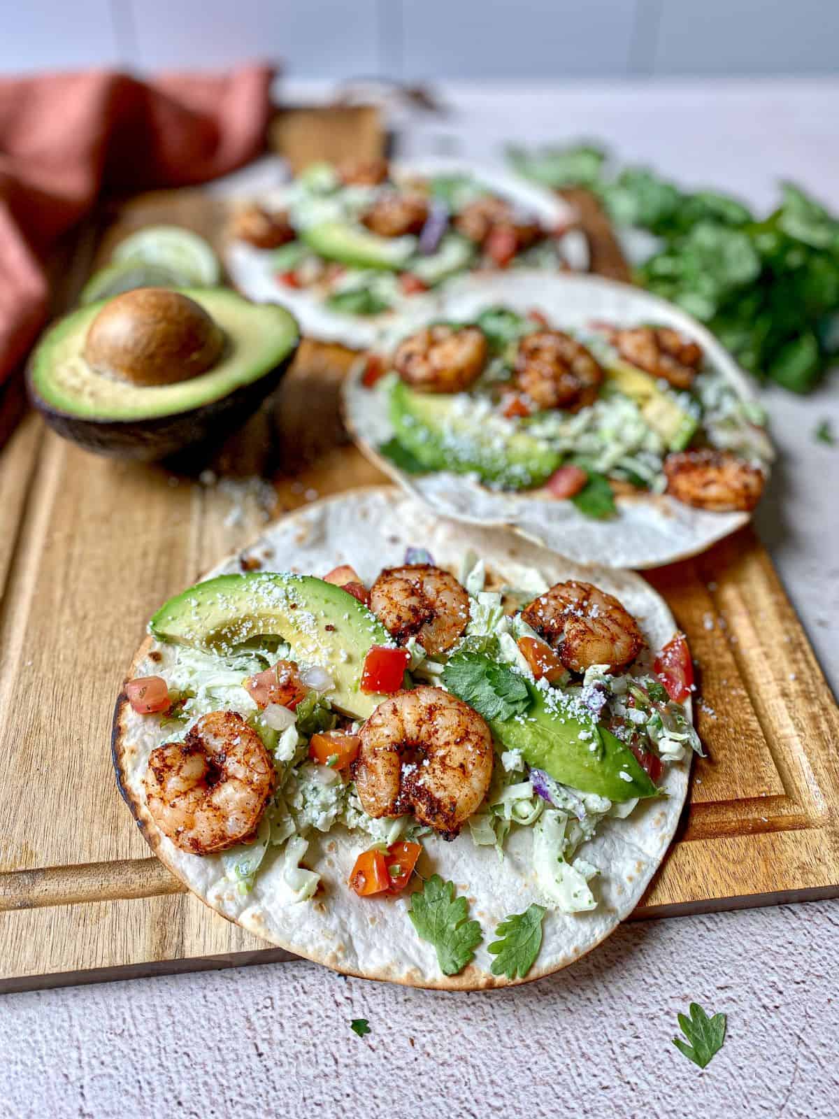 Shrimp tacos cooked in an air fryer are on a cutting board. Shrimp taco toppings are cabbage slaw, cilantro, avocado, and pico de gallo.