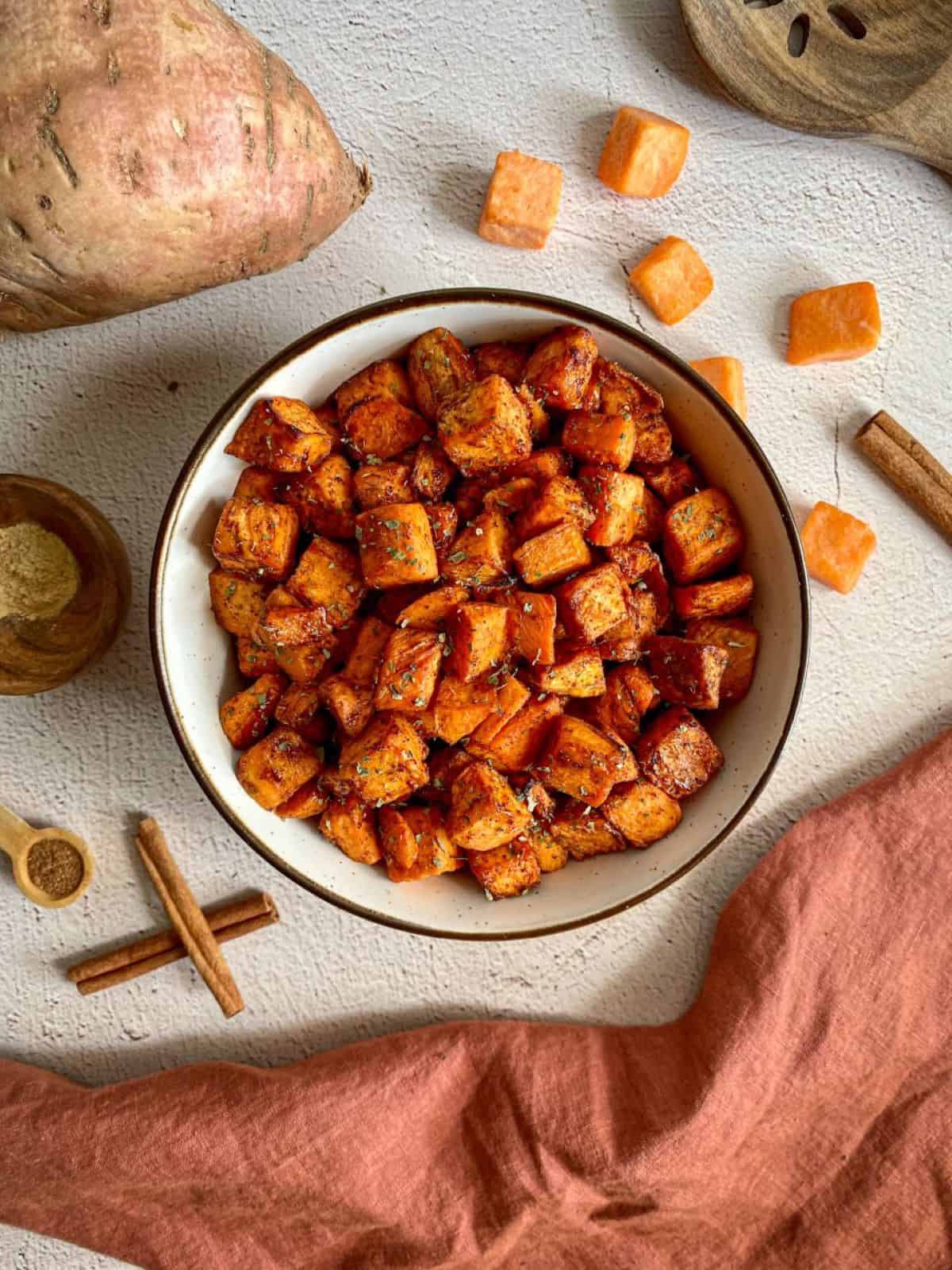 A bowl of air fried sweet potato chunks. Whole and cubed sweet potatoes, cinnamon sticks, and measuring spoons with nutmeg and ginger surround the bowl. 