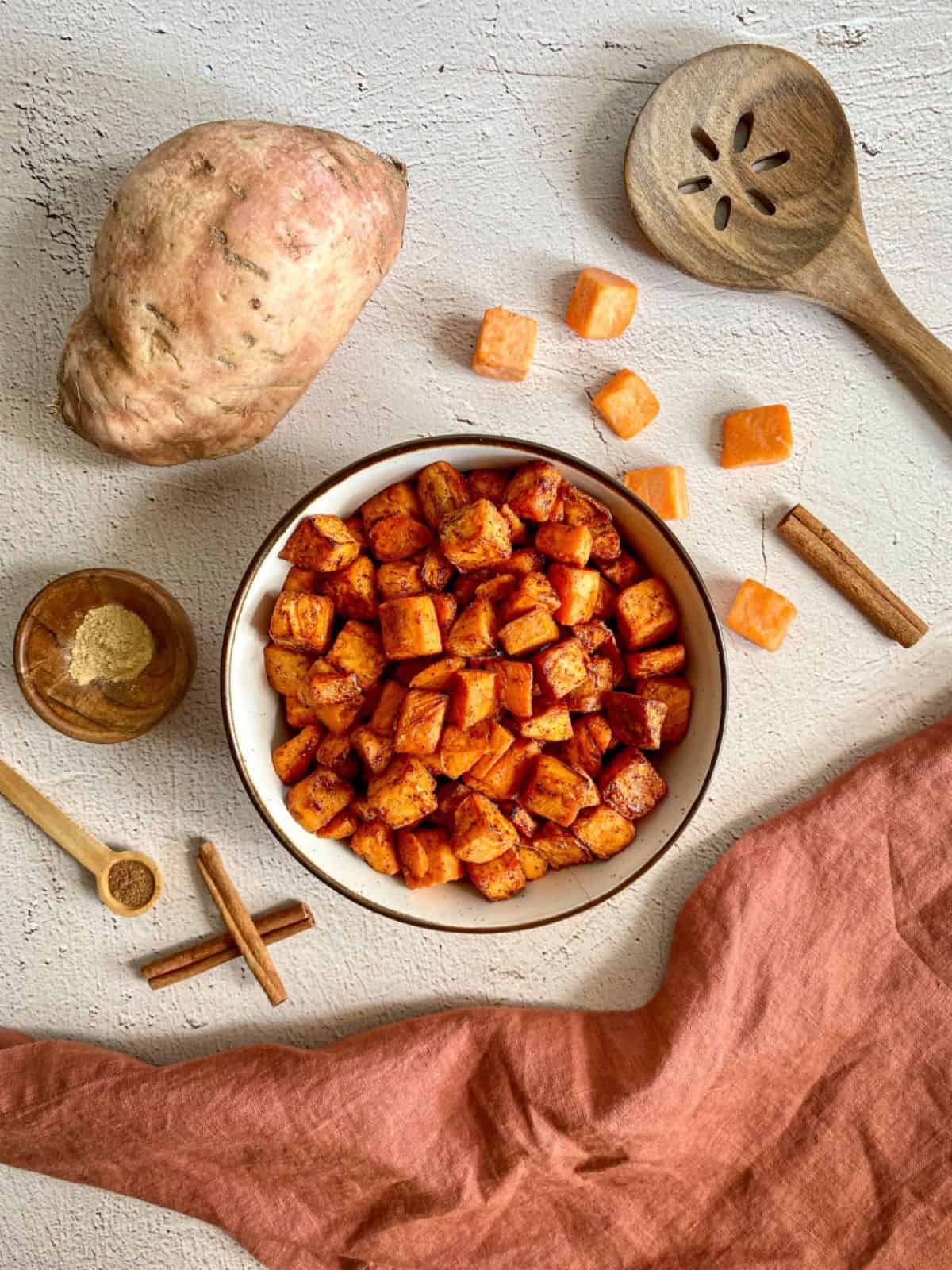 A bowl of air fried sweet potato chunks. Whole and cubed sweet potatoes, cinnamon sticks, and measuring spoons with nutmeg and ginger surround the bowl. A wooden spoon and rust colored towel are on the edges.