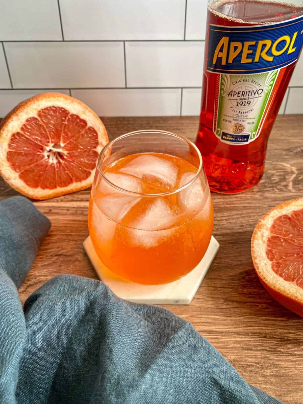 A glass of an orange colored cocktail with gin and aperol. A bottle of aperol and a sliced grapefruit are on the side of the cocktail with a blue cloth.