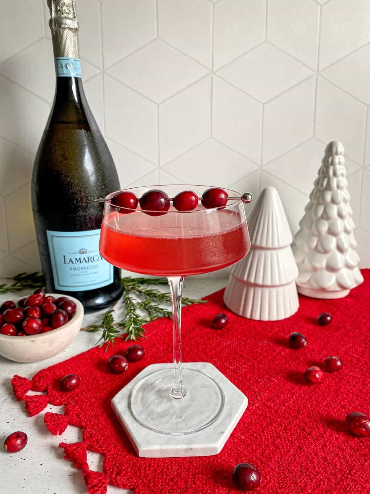 Cranberry gin cocktail sitting on top of a red placemat with a cranberry garnish. A bottle of prosecco and a bowl of fresh cranberries are behind the drink.