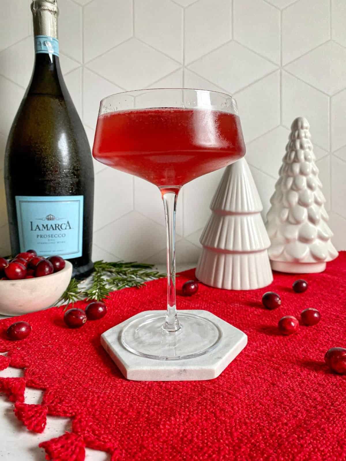Red cocktail in a coupe glass on top of a red placemat. A bottle of prosecco is in the background.