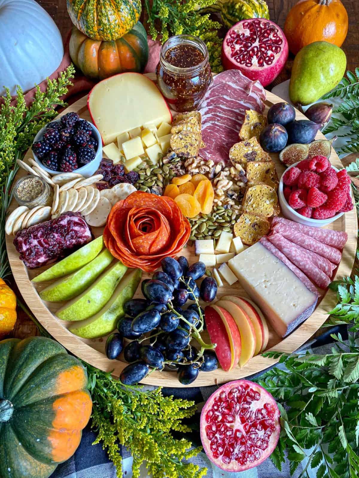 A fall themed charcuterie board full of cheeses, meats, and fruits. Greenery and fresh pumpkins surround the wooden board.