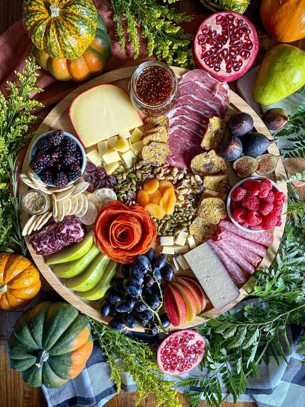 A fall charcuterie board arranged with three different types of cheese and variety of fruit and meat are on a round wooden board. Greenery and fresh pumpkins surround the board.