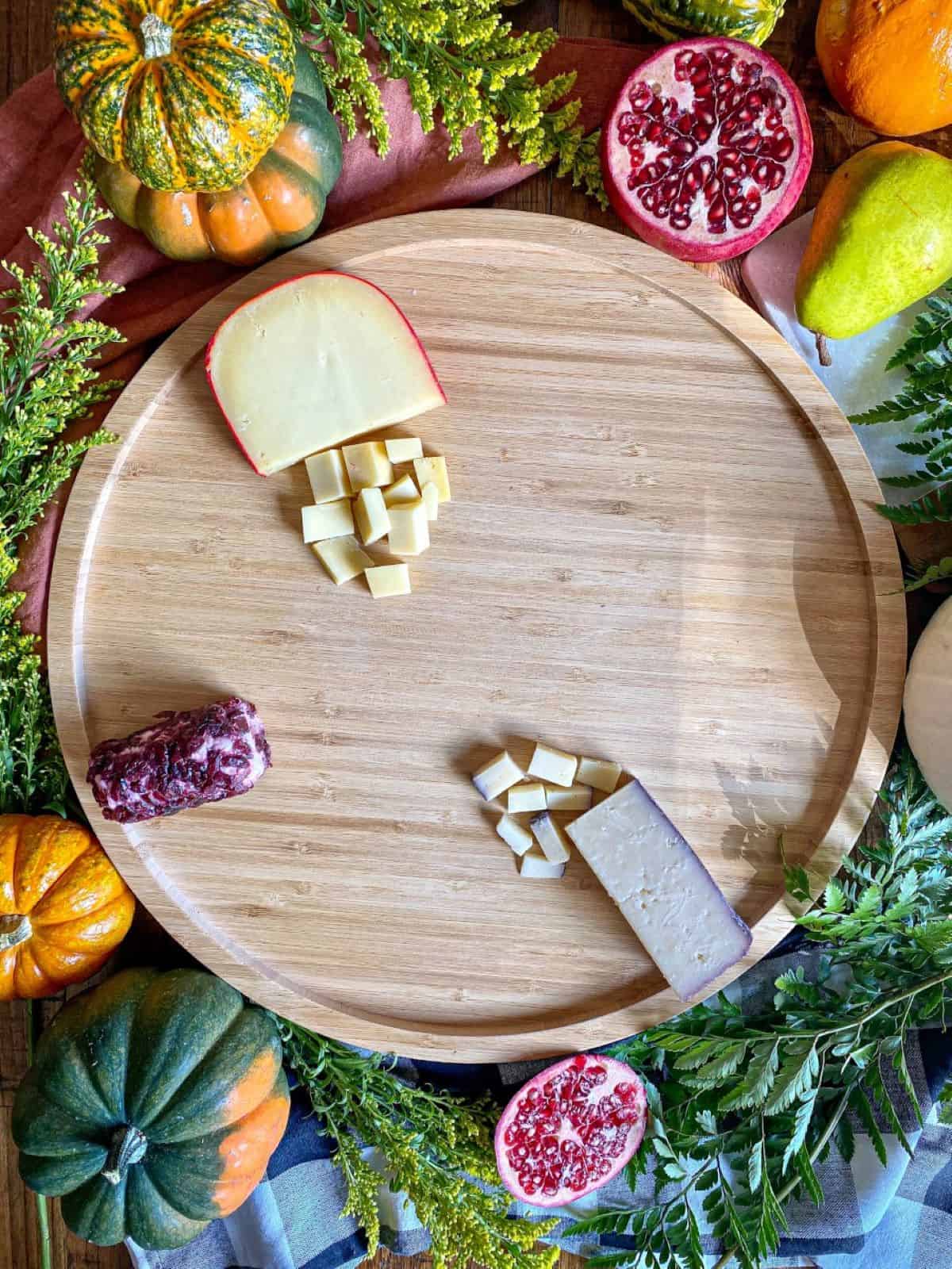 Three different types of cheese are on a round wooden board. Greenery and fresh pumpkins surround the board.