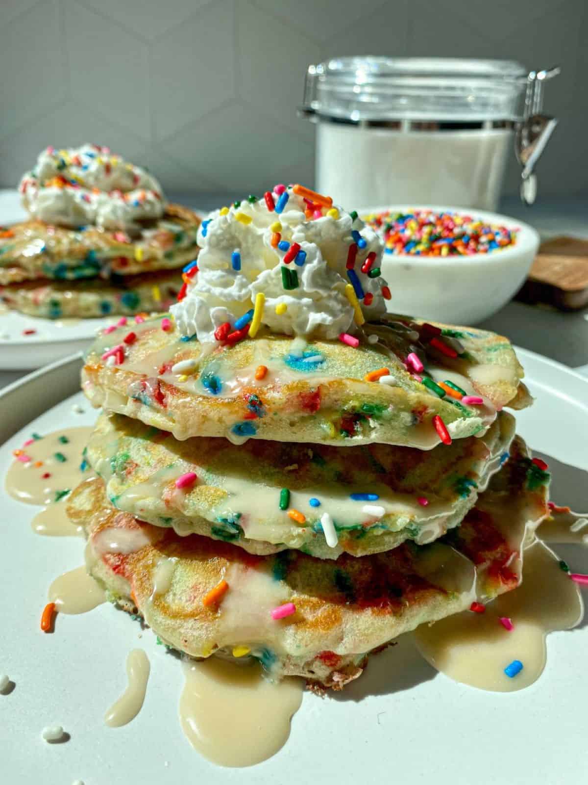 Two stacks of  homemade funfetti pancakes with sprinkles on white plates with a bowl of rainbow sprinkles on the side.