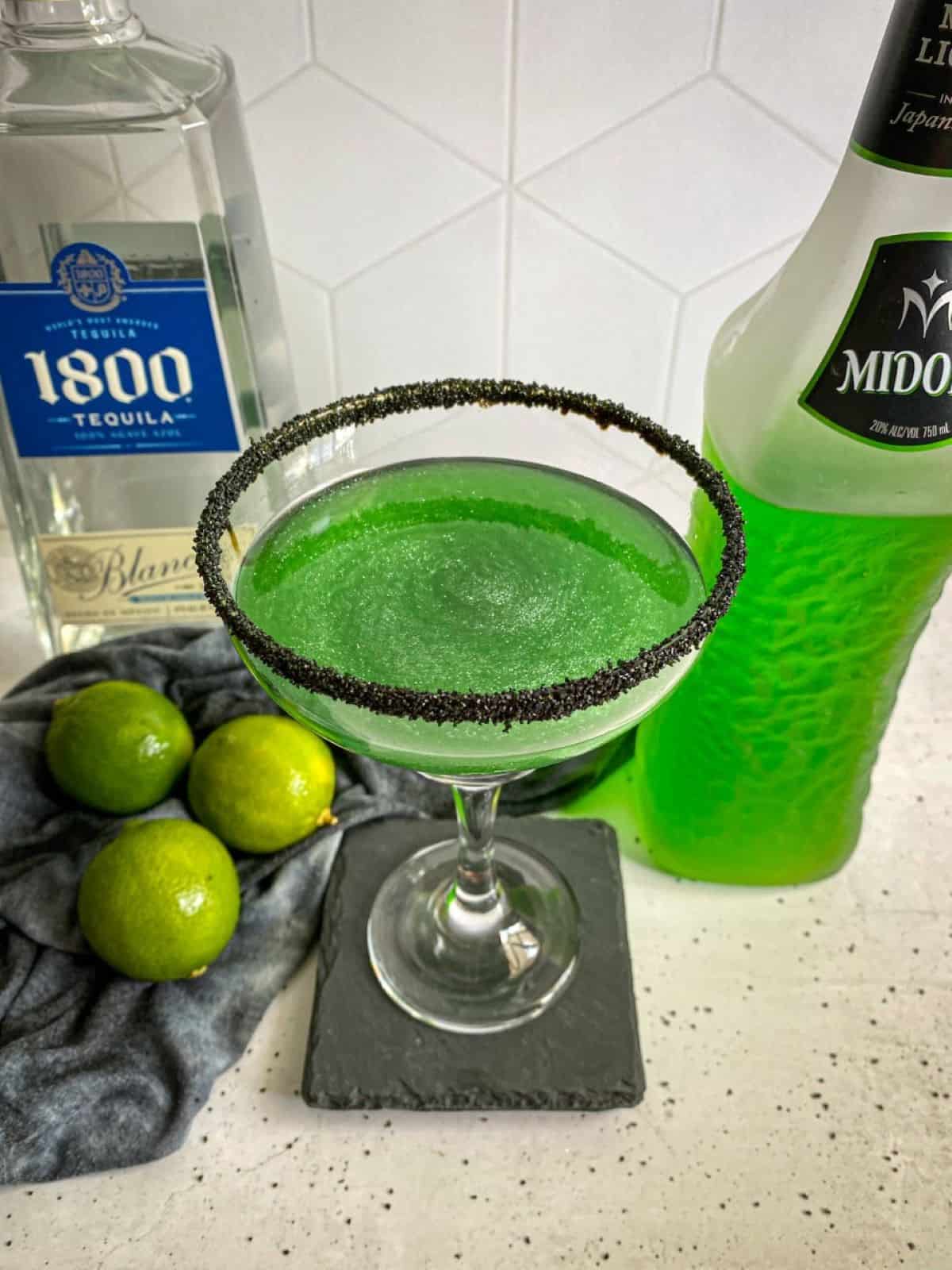 Halloween margarita with a black sugar rim. A bottle of midori melon liqueur and tequila are in the background with three limes.