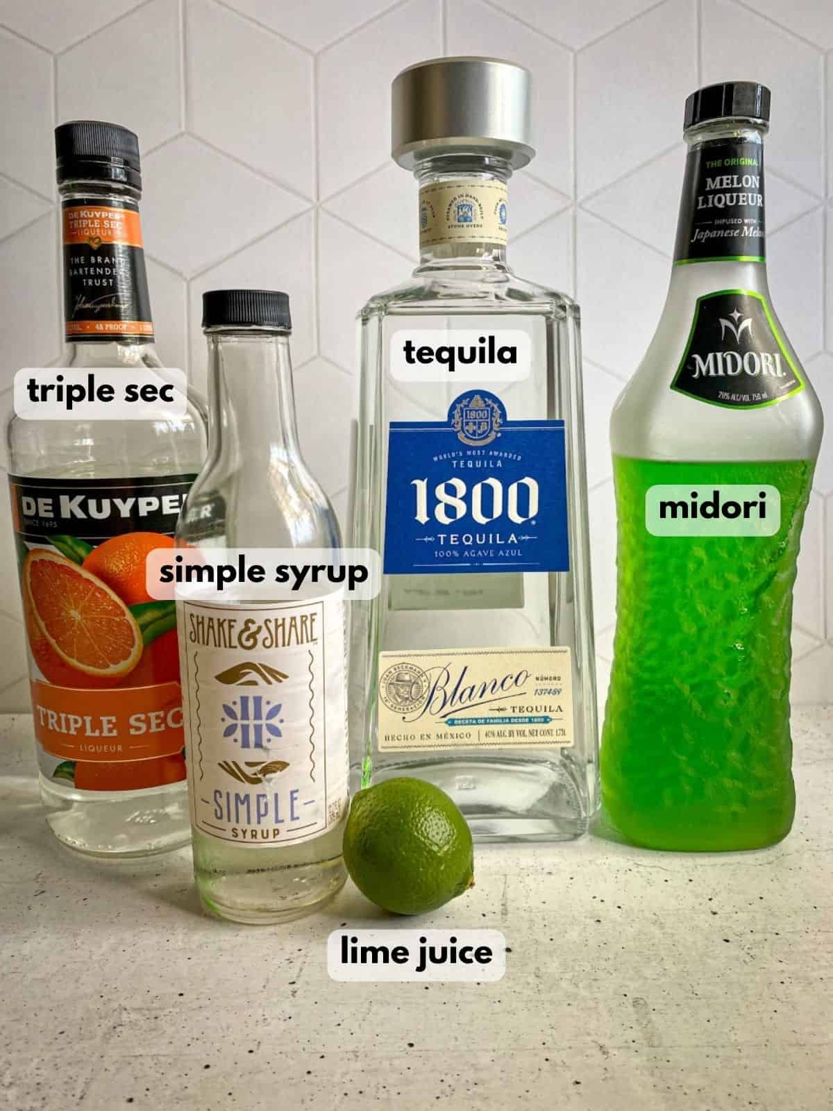 Ingredients needed to make witches brew Halloween margarita. A fresh lime, bottles of tequila, midori, triple sec, and simple syrup are on a concrete table.