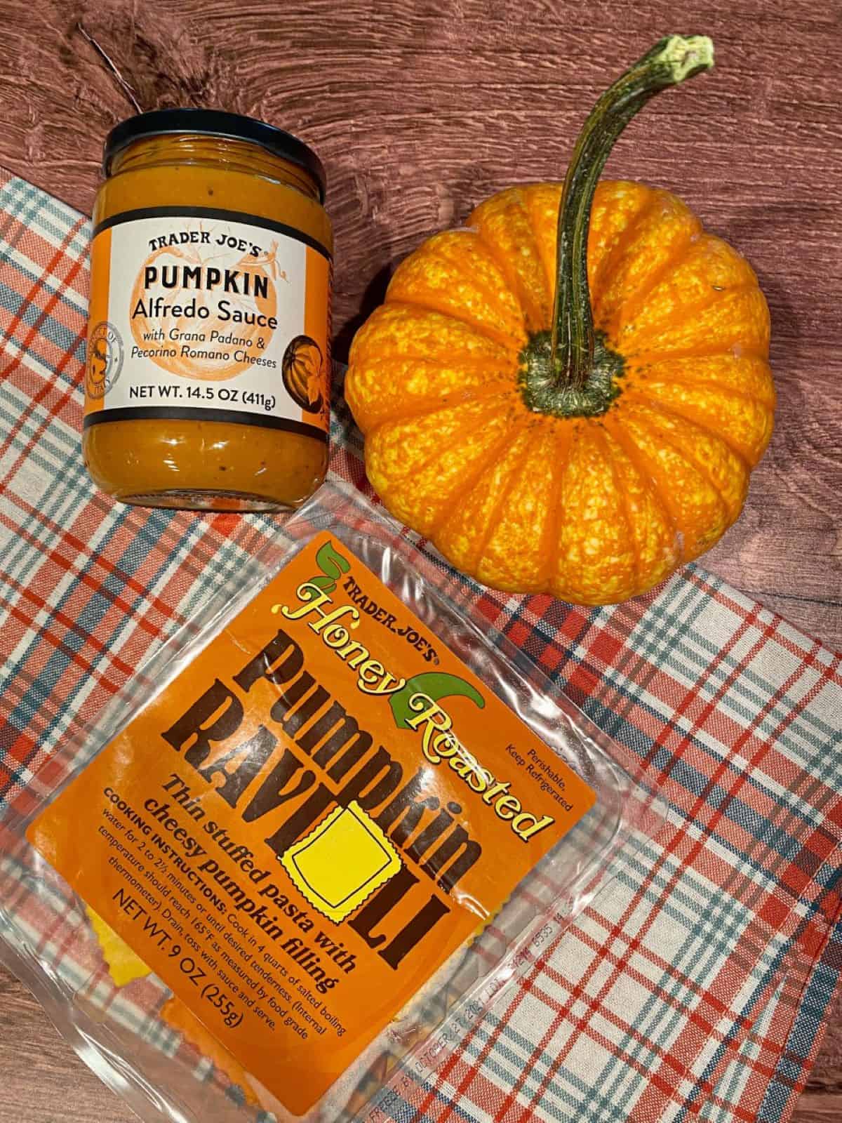 Ingredients needed to make this recipe, pumpkin ravioli and a jar of alfredo sauce, are on a wooden table with an orange and blue fall towel.