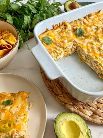 Sour Cream Chicken Enchilada Casserole cut into with one serving of the layered chicken casserole on a plate. Cilantro, avocado, and chips surround the meal.