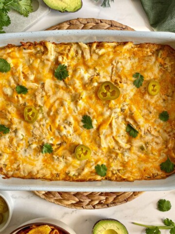 Sour Cream Chicken Enchilada Casserole in a large baking dish. A green kitchen towel, cilantro, avocado, and chips are around the dish.