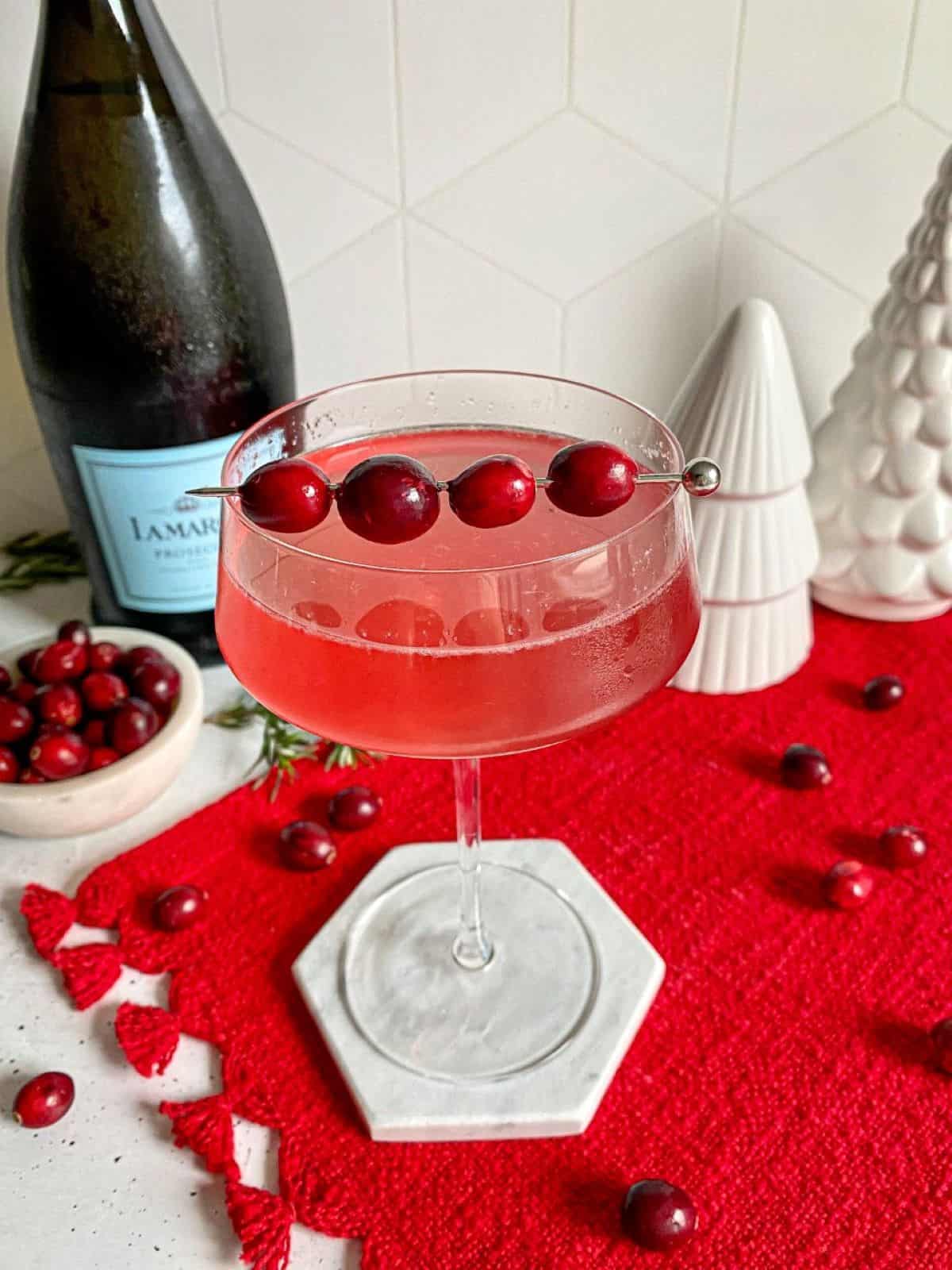 Red Cocktail in a coupe glass with a garnish of fresh cranberries on top of a red placemat. A bottle of prosecco and fake white Christmas tree decorations are in the background.