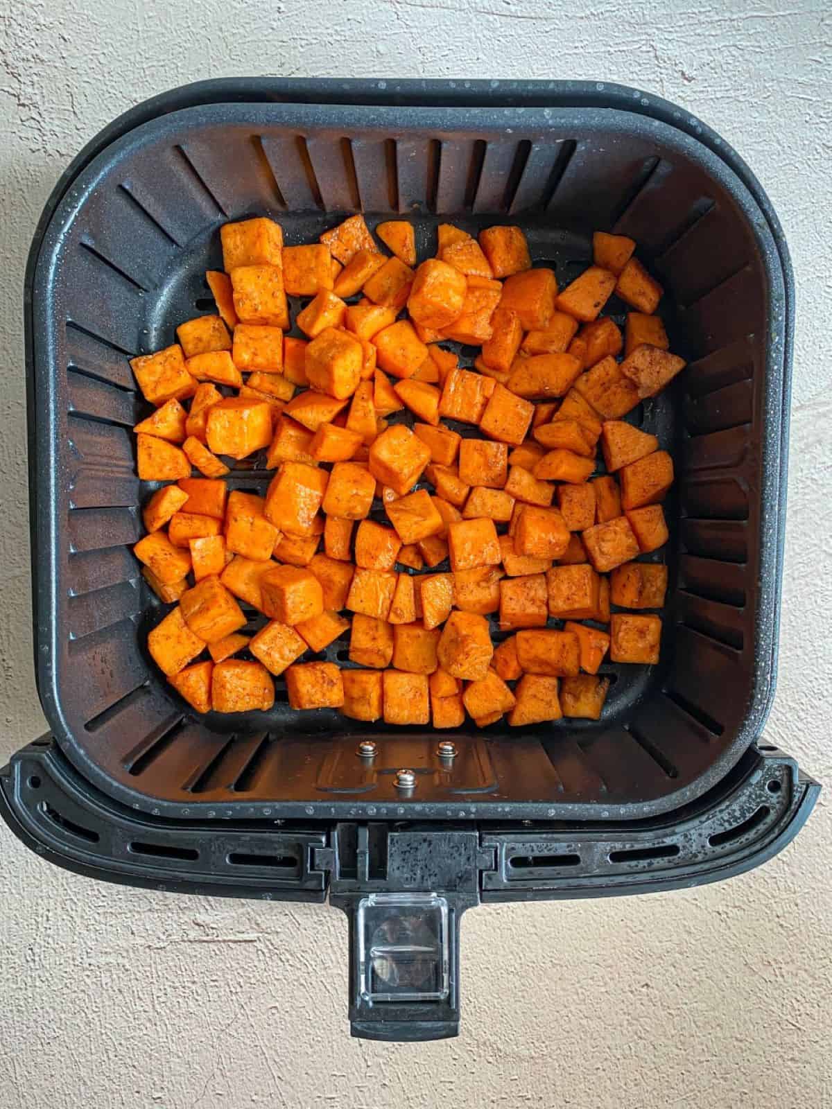 Diced and seasoned sweet potato are in a single layer in an air fryer basket.