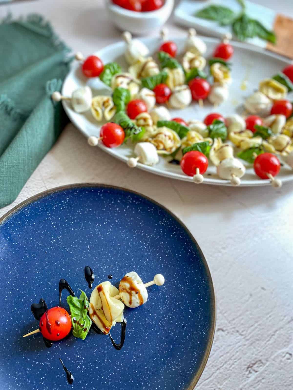 One caprese skewer is on a blue plate. Multiple tortellini skewers with balsamic glaze are on a serving tray in the background. 