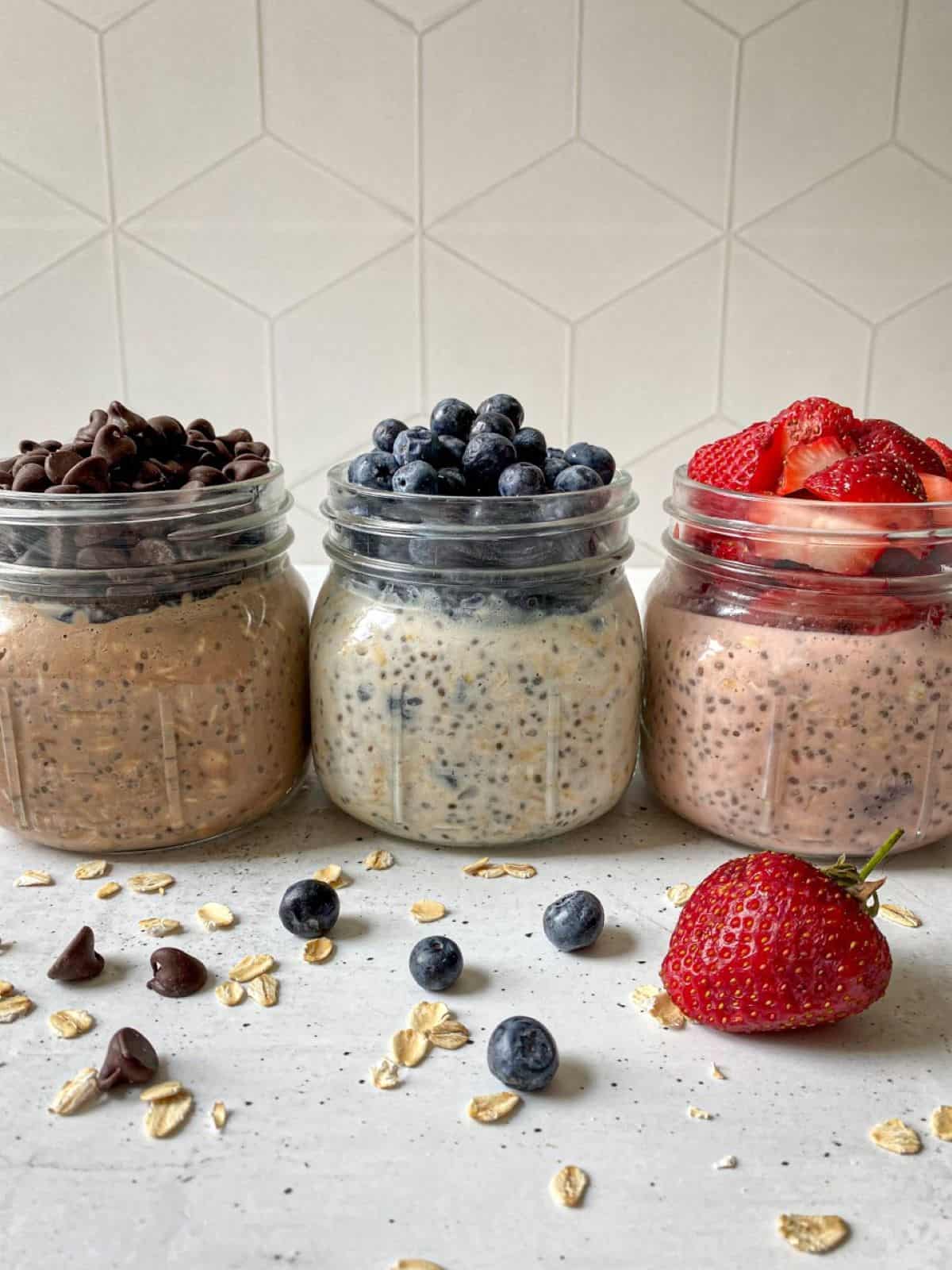 Three mason jars filled with high protein overnight oats. The three flavors include: Chocolate chip overnight oats have chocolate chips on top, blueberry cheesecake overnight oats has blueberries on tope, and strawberry shortcake overnight oats has chopped strawberries on top.
