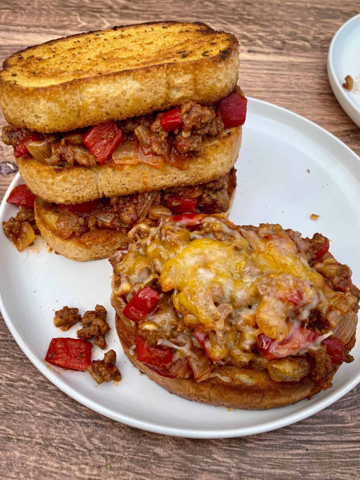 Open faced sloppy joes with cheese and a stacked sloppy joes on Texas toast. Both sloppy joes sandwiches are on a white plate.