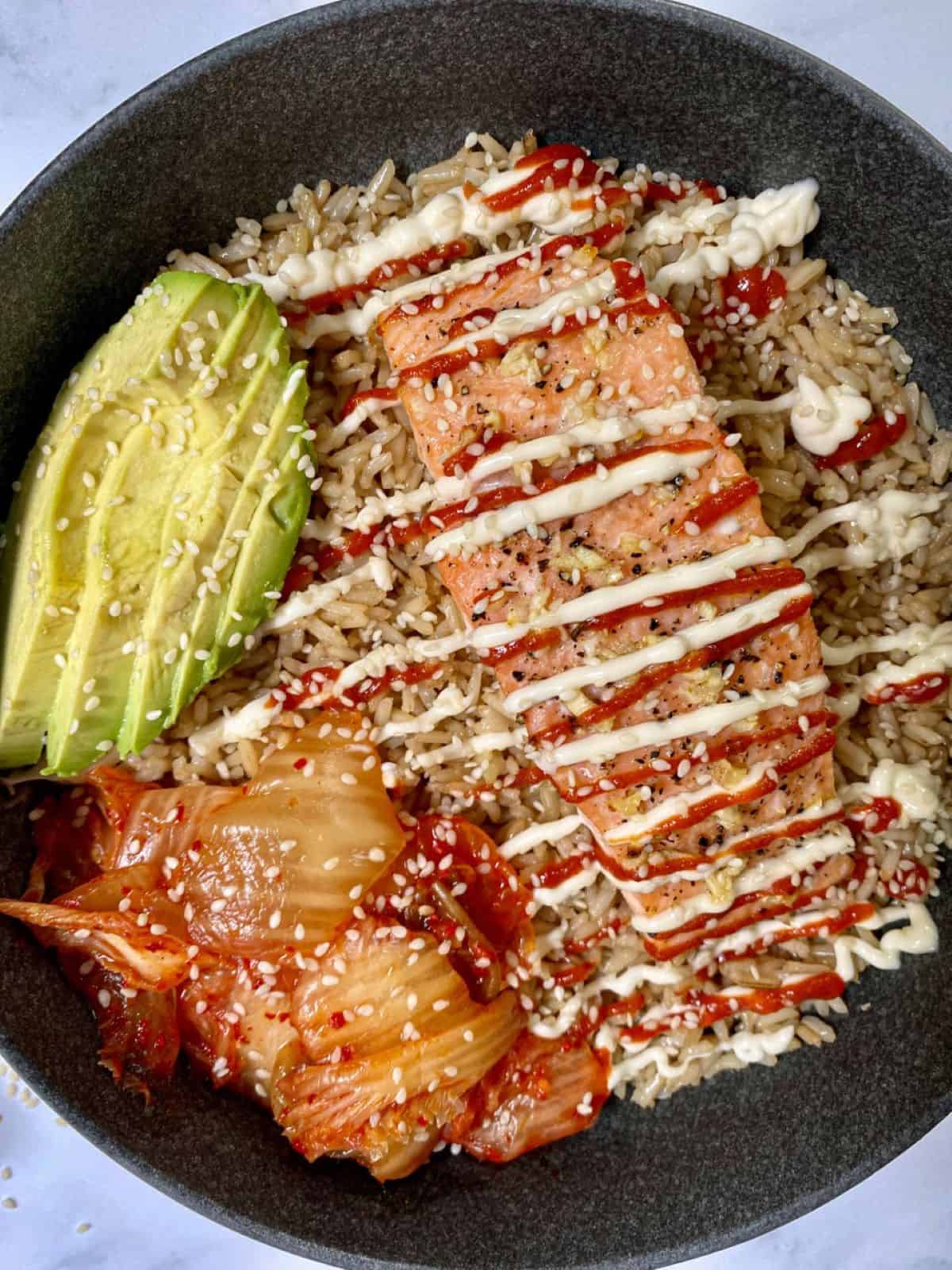A bowl with salmon, rice, kimchi and avocado with a drizzle of mayo and siracha sauce.