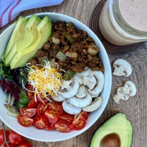 Beef taco salad topped with cheese, tomatoes, sliced avocado, and mushrooms in a bowl with a jar of salsa ranch dressing on the side.