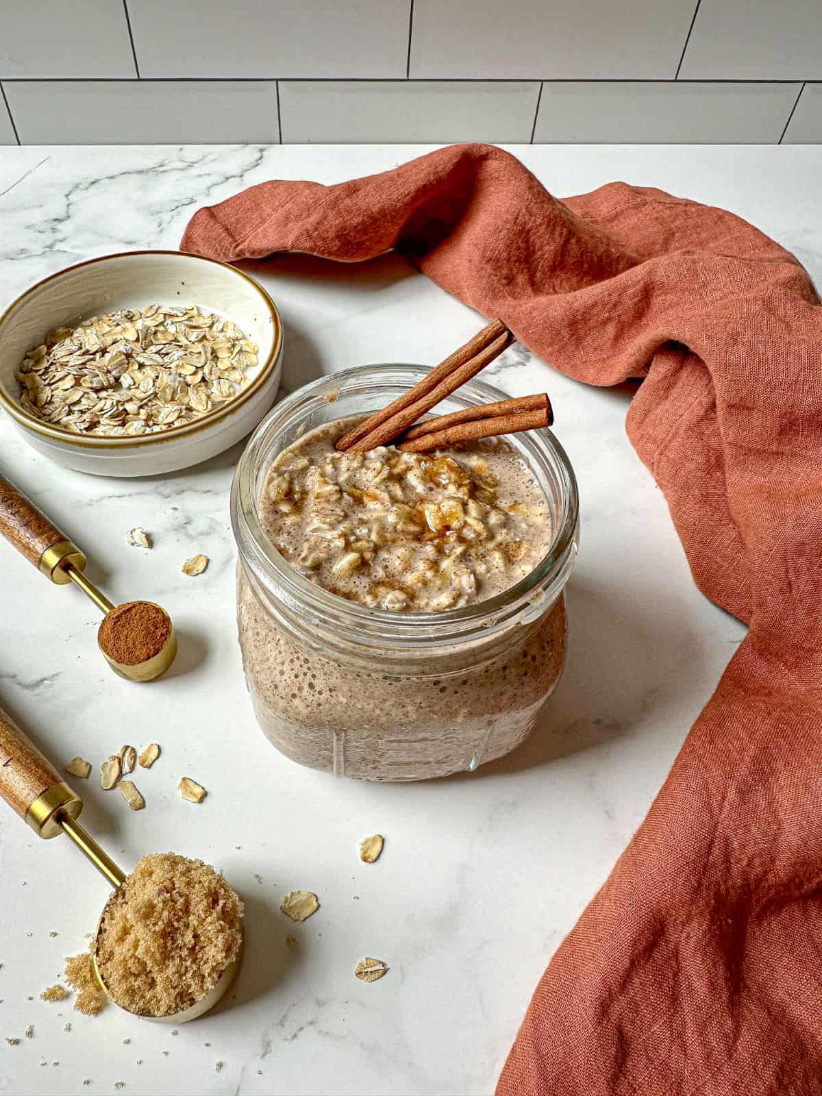 A mason jar filled with cinnamon overnight oats are on a marble countertop. Ingredients like a bowl of oats, cinnamon, and brown sugar sit on the counter next to the jar.