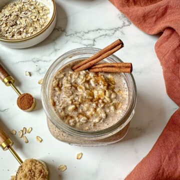 Cinnamon roll overnight oats in a mason jar with two cinnamon sticks. Measuring spoons with brown sugar and cinnamon, and a bowl of oats are on the side.