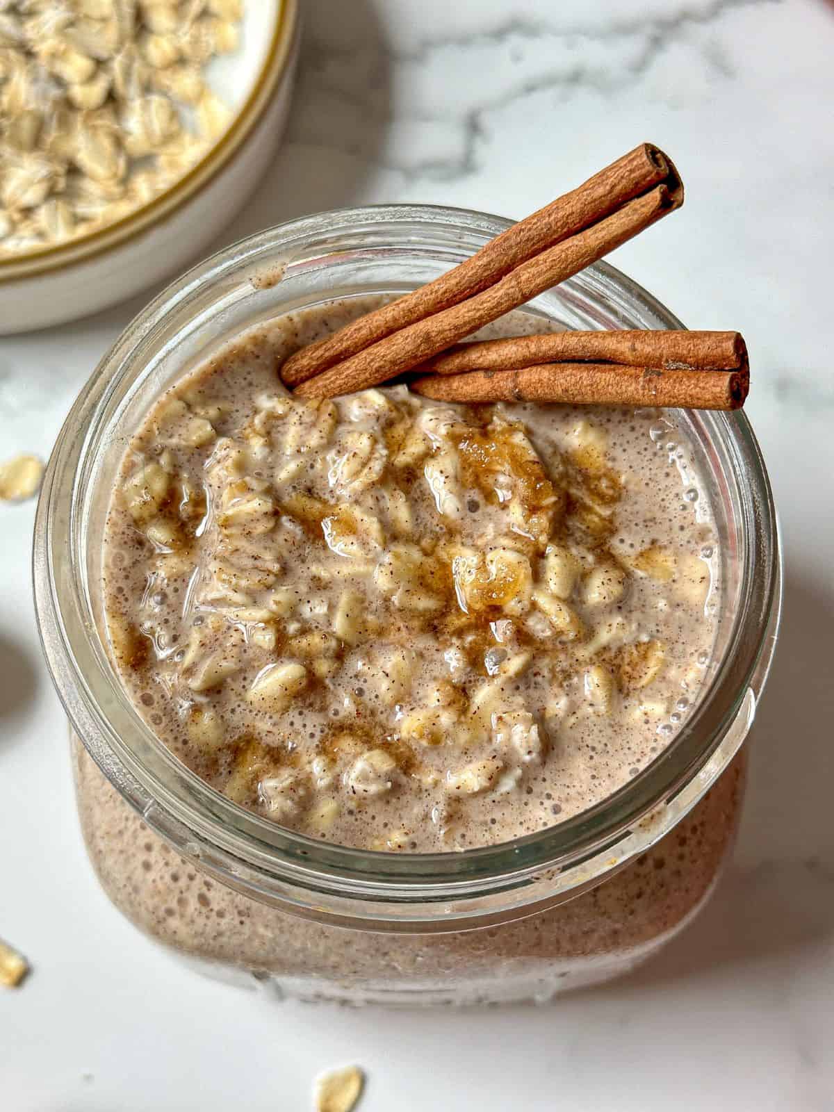 Mason jar full of overnight oats with cinnamon and brown sugar. Two cinnamon sticks are on top of the oats.