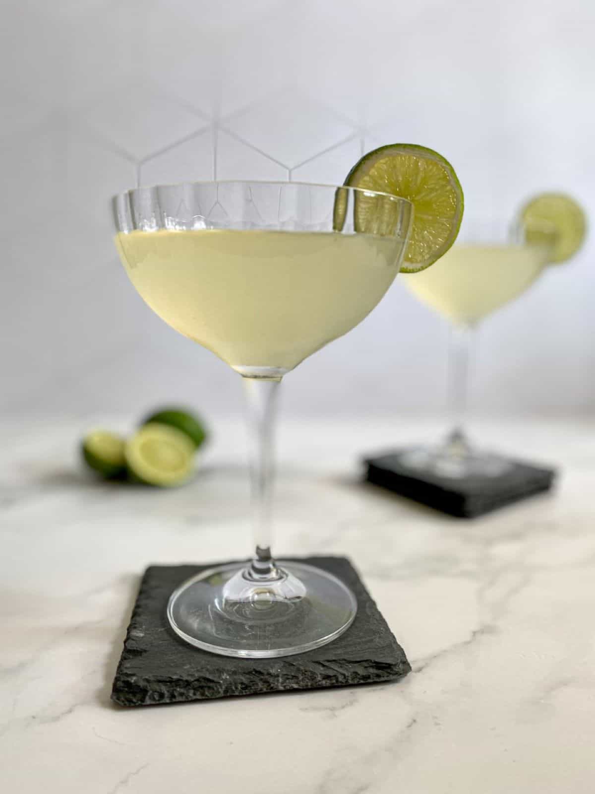 French gimlet cocktail with a lime wedge garnish is in the forefront with a second cocktail and lime wedges in the background.