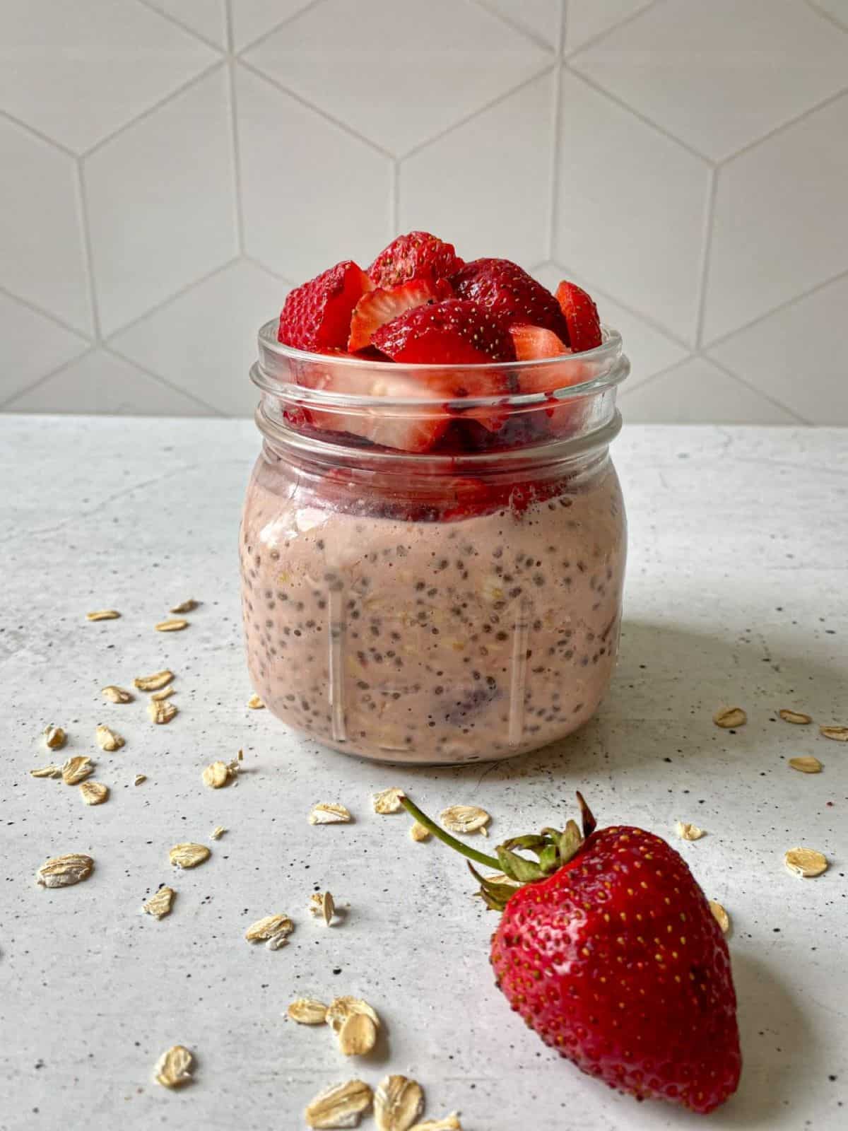 Strawberry Overnight Oats in a mason jar topped with chopped fresh strawberries. A large strawberry and oats are scattered around the jar.