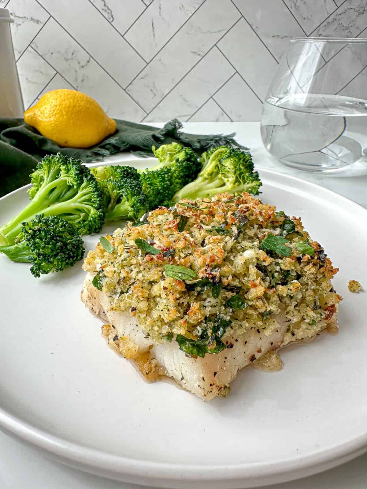 Parmesan panko crusted cod on a plate with broccoli. A green towel, glass of water, and lemon are in the background.