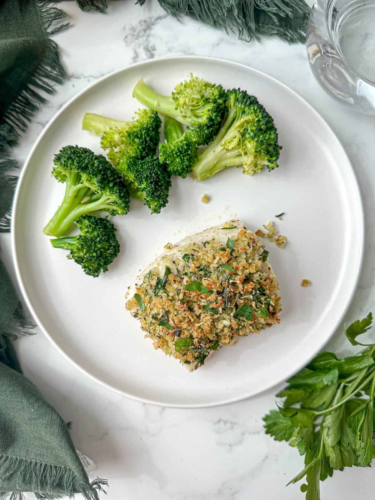 Baked parmesan crusted cod on a white plate with broccoli. Crust is golden brown and sprinkled with green parsley.