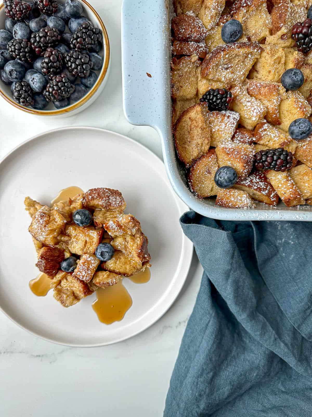 Baked French toast casserole in a baking dish. A plate with a square slice is next the dish. The brioche casserole is topped with blueberries, blackberries, and powdered sugar.