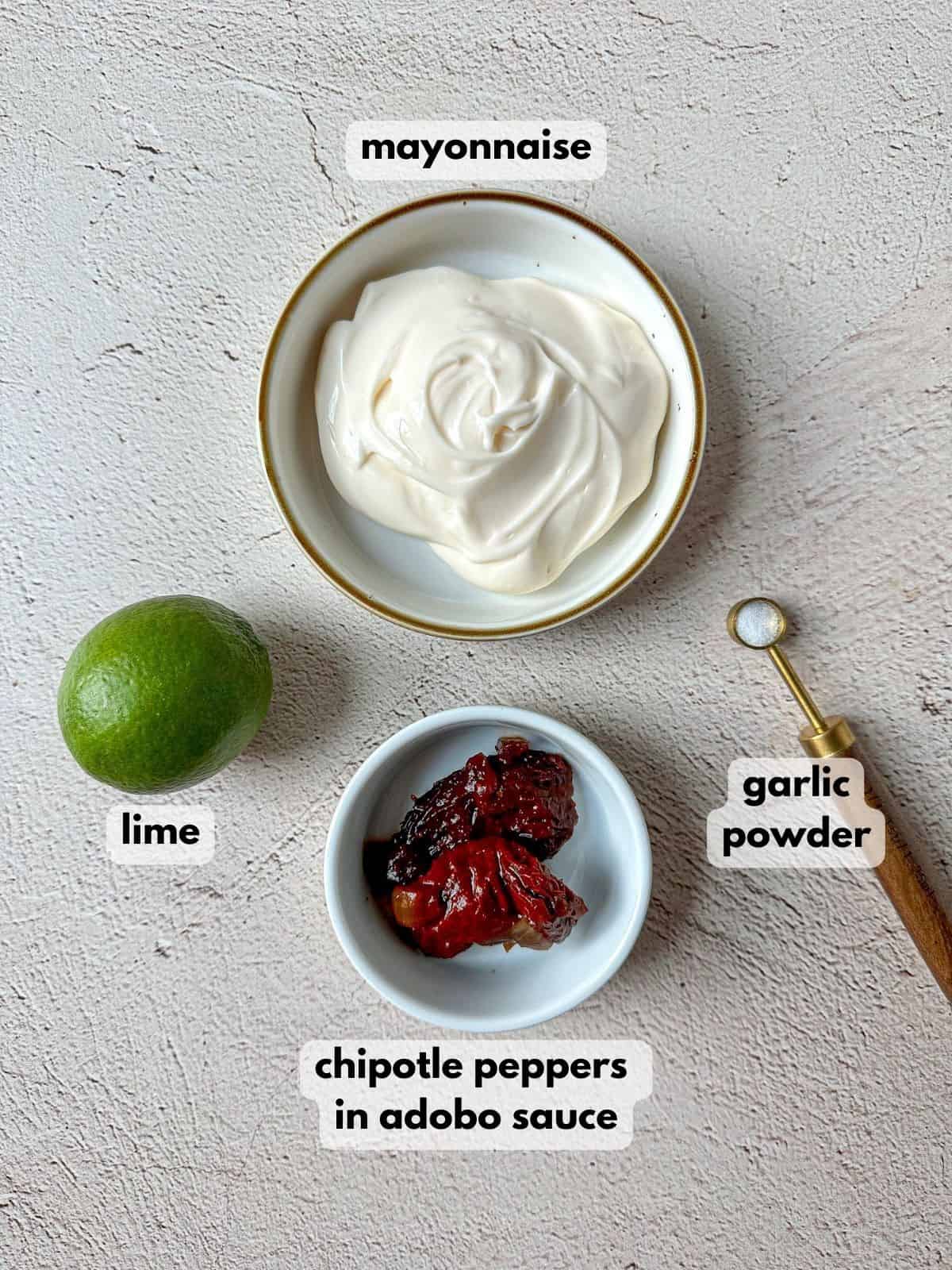Ingredients needed to make this spicy sauce on a textured table: mayonnaise, whole lime, chipotle peppers in adobo sauce, and a measuring spoon of garlic powder. 