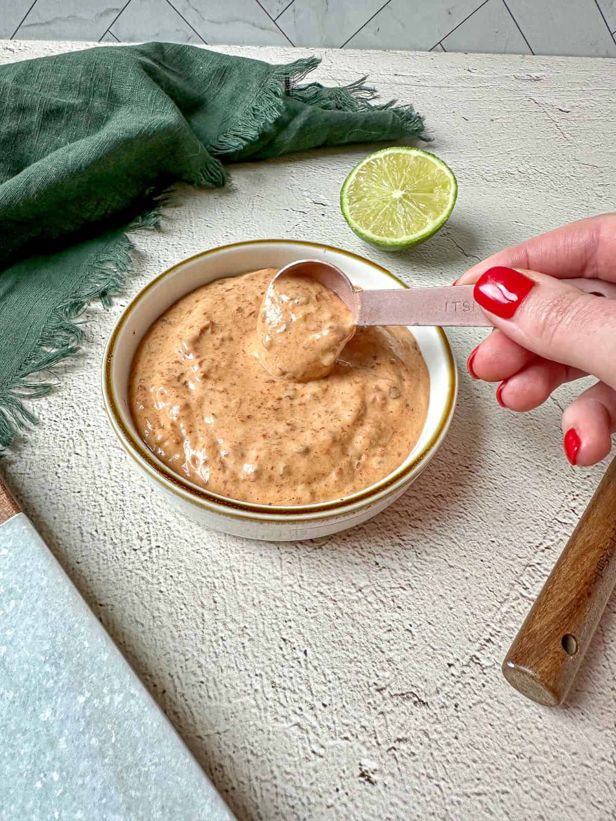 A hand holding a measuring spoon dipping into a bowl of the light orange colored mayo sauce. Sauce is smooth with a few chunks of chipotle peppers.