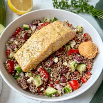 This salmon quinoa bowl recipe in a white bowl. A Greek salmon fillet is on top of a quinoa salad tossed with vinaigrette and fresh produce.