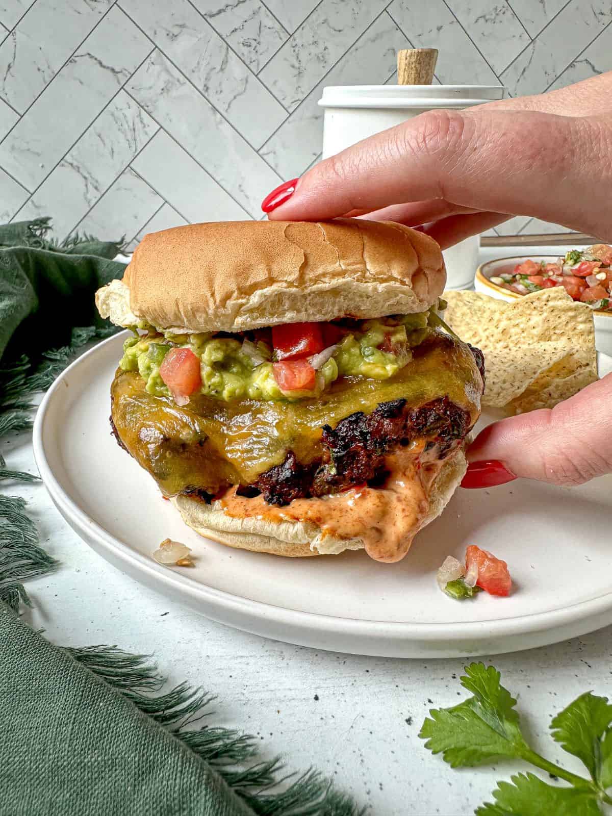 A hand with red nails is grabbing a taco burger topped with guacamole, pico de gallo, and spicy mayo dipping out from the bottom bun.
