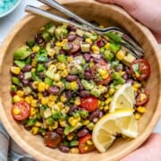 Two hands holding a wooden bowl filled with cowboy caviar with black beans, corn, and cherry tomatoes.