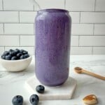 Blueberry protein shake in a clear glass with a straw. Blueberries, chia seeds, and nuts surround the glass.