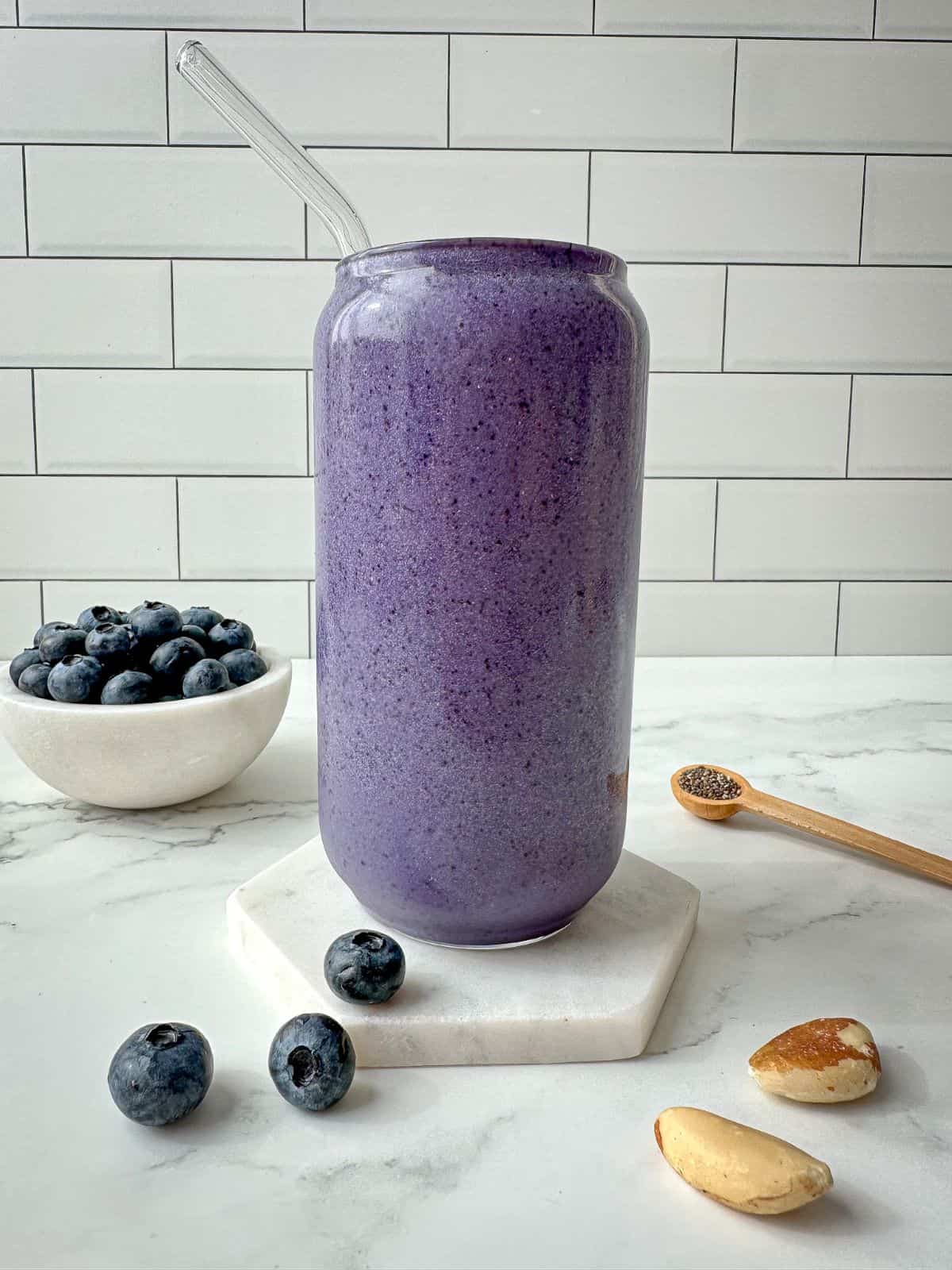 Blueberry protein shake in a clear glass with a straw. Blueberries, chia seeds, and nuts surround the glass.