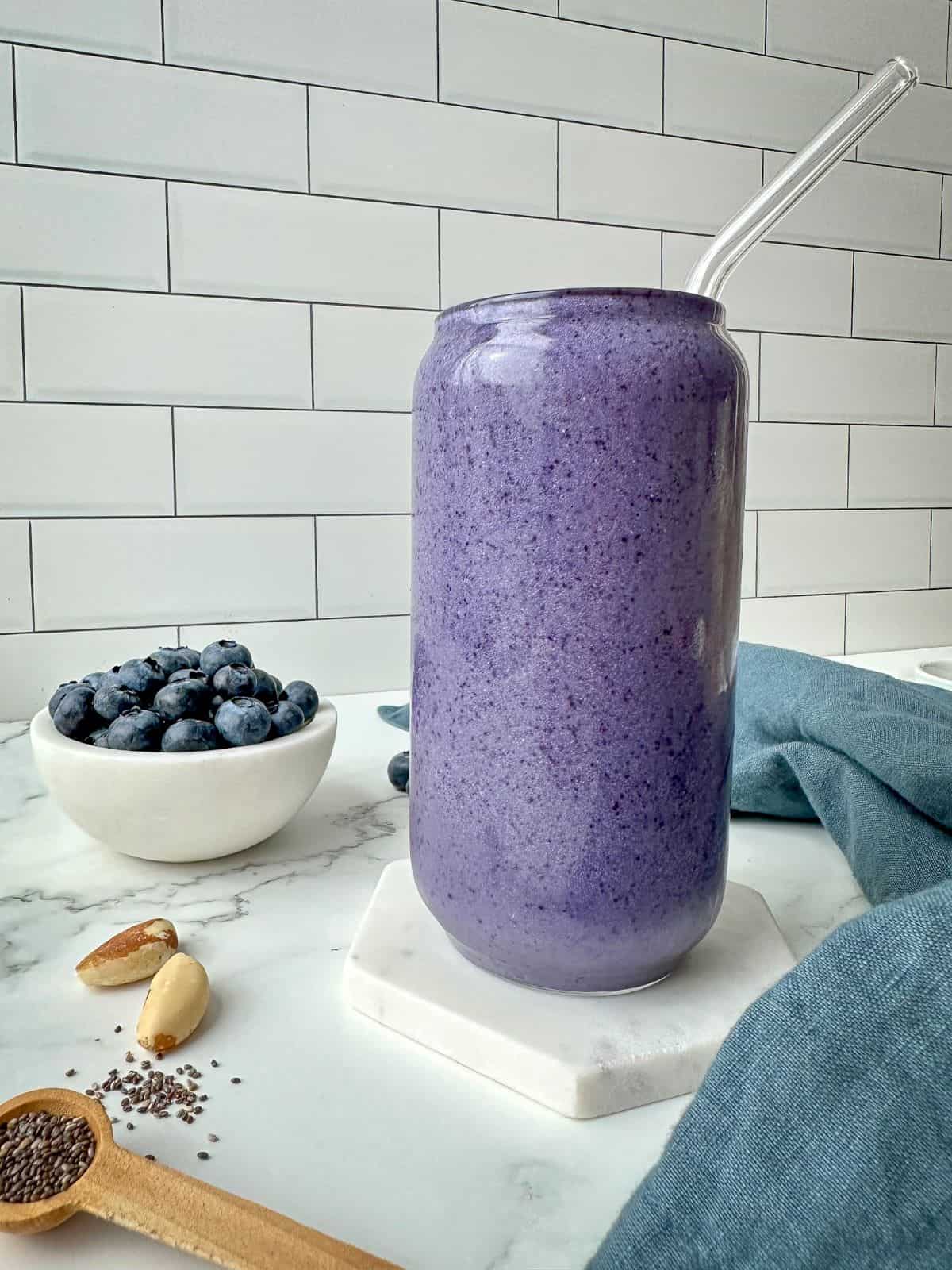A purple smoothie in a clear glass. A bowl of blueberries, Brazil nuts, and chia seeds are on a marble table with a blue kitchen towel.