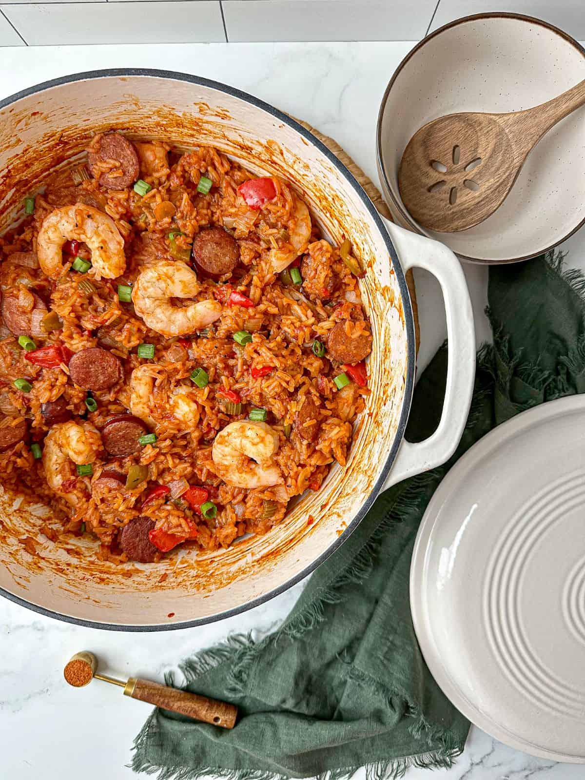 Red creole jambalaya cooked in a Dutch oven. The oven lid and a white bowl with a wooden spoon are next to the pot.